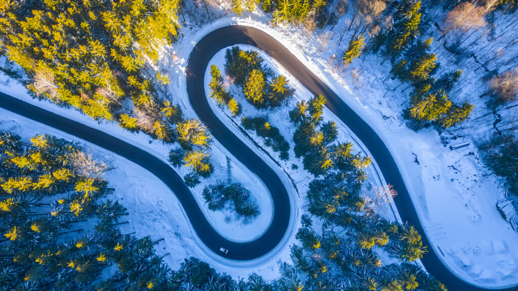 Asphaltstraße Serpentine im verschneiten Winter. Kalter Winter und sonniger Tag über der Forststraße mit beleuchteten Bäumen bei aufgehender Sonne. Luftaufnahme von Drohne.