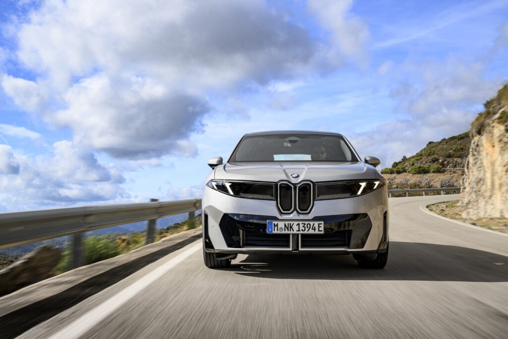Grauer BMW ix3 fährt auf einer Straße mit blauem Himmel und leichter Wolke