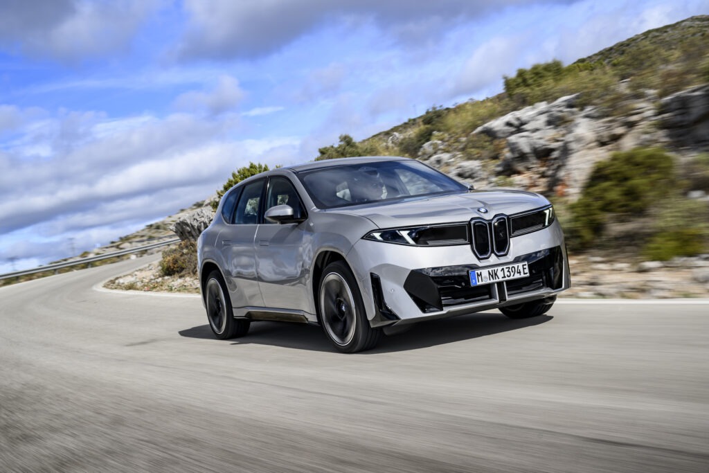 Grauer BMW ix3 fährt auf einer Straße mit blauem Himmel und leichter Wolke