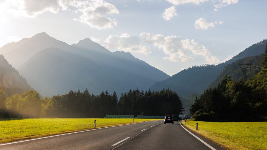 Scenic back view cars traffic driving stunning alpine road warm sunset sunrise light sky background mountain silhouette. Landscape curvy winding vehicle freeway route evening sunlight.