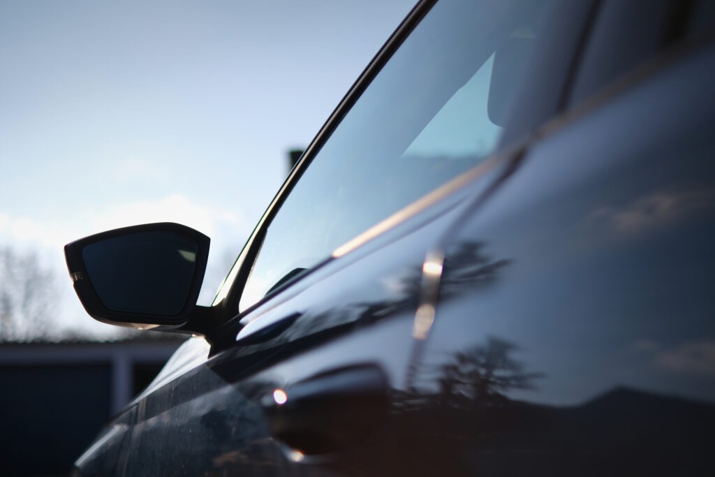 Close-up of a car’s side mirror and door with glossy reflections of the sky and trees, showcasing a sleek design, smooth curves, and tinted windows.